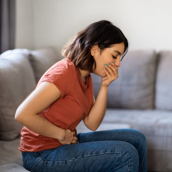 Food Poisoning Concept. Young Arab Female Feeling Sick At Home, Ill Middle Eastern Woman Suffering From Nausea, Covering Mouth And Touching Belly While Sitting On Couch In Living Room, Side View